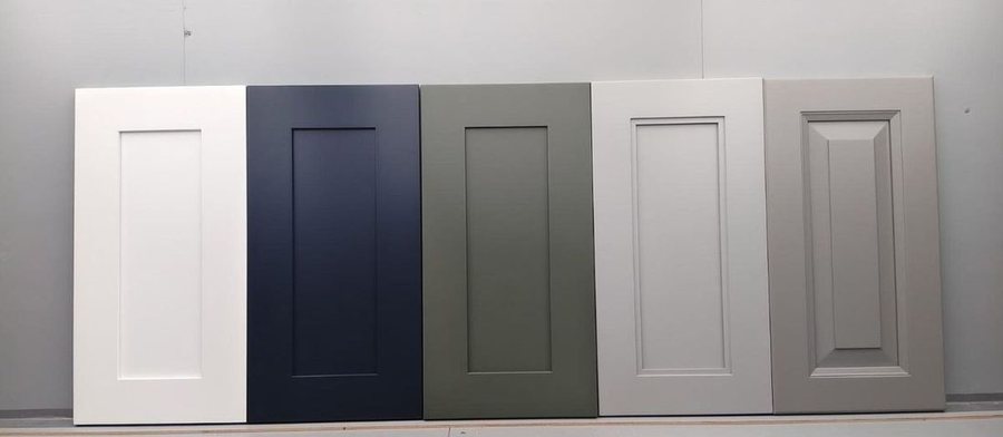 Shaker cabinet door samples in white, navy, sage and light grey, with a warm grey raised panel door, leaning against a wall